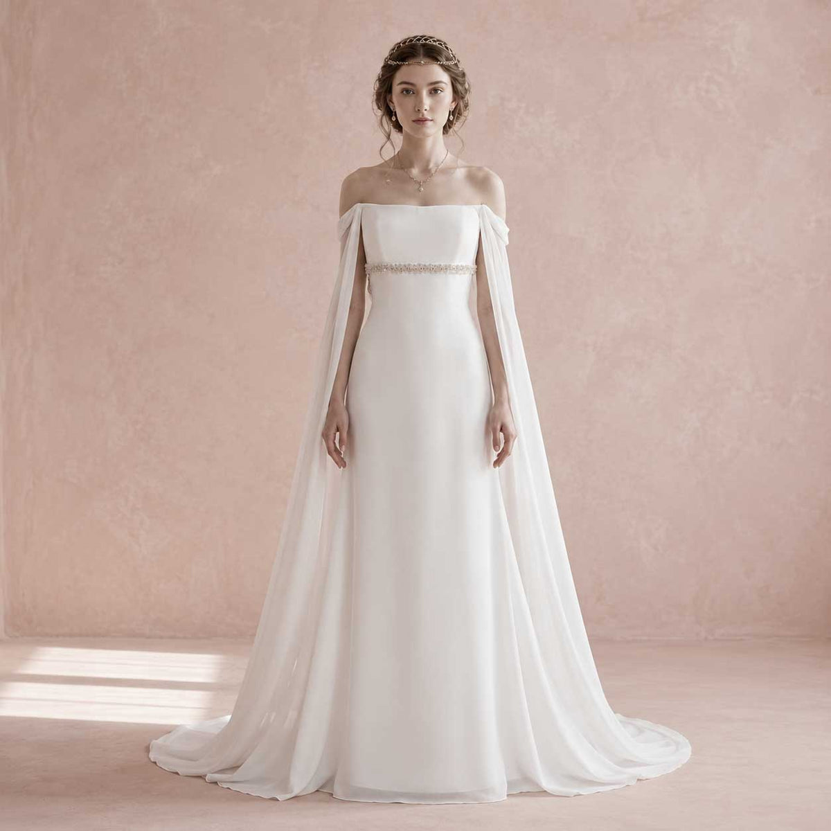 A woman modeling an off-the-shoulder white wedding gown with a flowing sheer cape and a thin beaded waistband, standing against a soft pink textured studio wall.