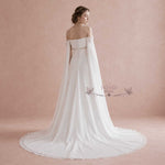 A bride wearing an off-the-shoulder white wedding gown with a long flowing train, photographed from the back against a soft pink textured studio backdrop. The dress has lace-up detailing at the back and a sheer draped cape-like overlay.