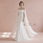 A woman models an off-the-shoulder white wedding gown with a fitted bodice, a thin embellished waistband, and a flowing sheer cape train against a soft pink studio backdrop. The brand name 'ieie BRIDAL' appears as a watermark on the dress.