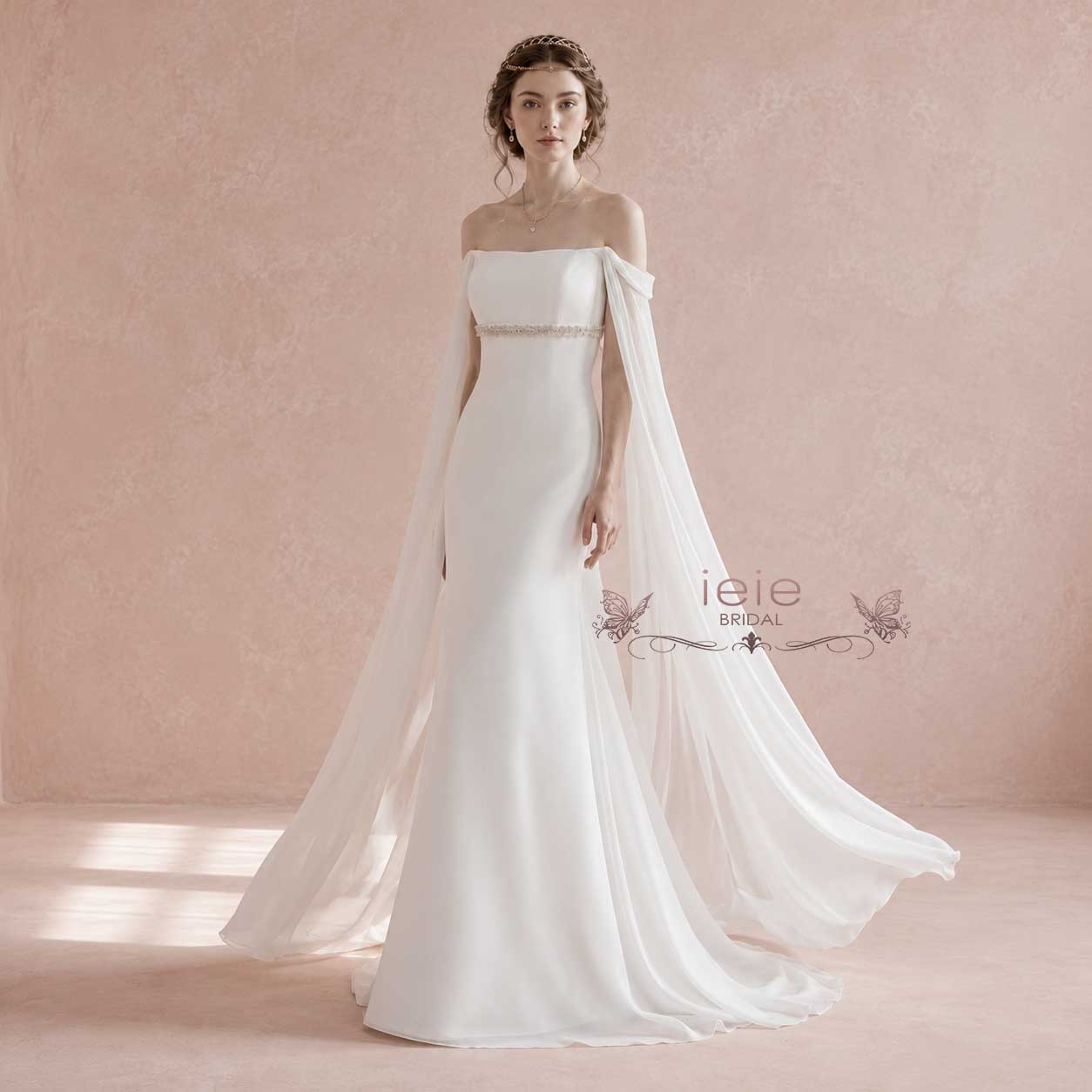 A woman models an off-the-shoulder white wedding gown with a fitted bodice, a thin embellished waistband, and a flowing sheer cape train against a soft pink studio backdrop. The brand name 'ieie BRIDAL' appears as a watermark on the dress.