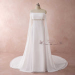 A white off-the-shoulder wedding gown with a sheer cape overlay and a beaded waistband, displayed on a mannequin against a textured pink studio backdrop.