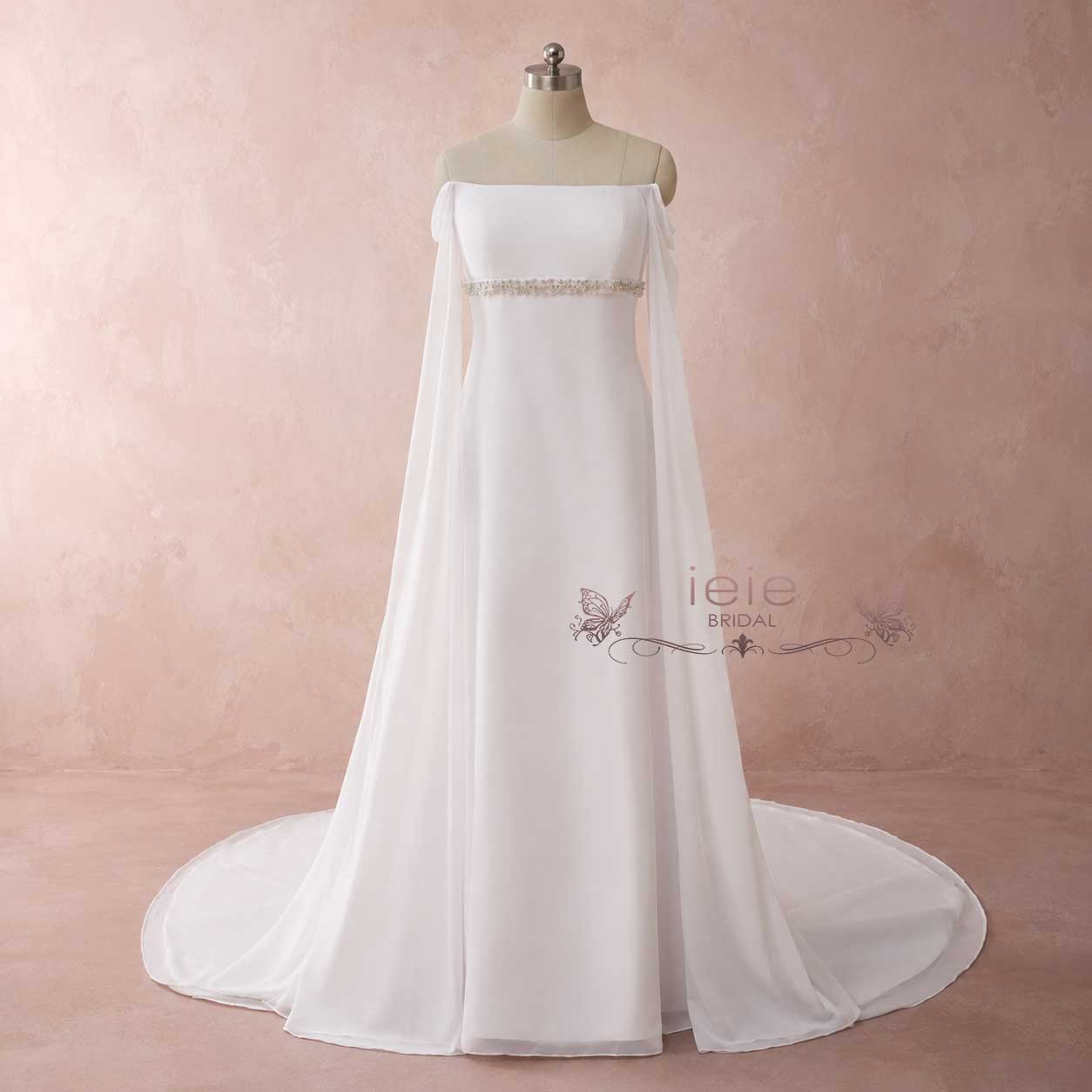A white off-the-shoulder wedding gown with a sheer cape overlay and a beaded waistband, displayed on a mannequin against a textured pink studio backdrop.