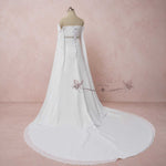 White strapless wedding dress with a long train displayed on a mannequin against a soft pink studio backdrop. The dress features a structured bodice, a central lace-up back detail, and a sheer draped overlay.