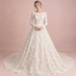 A woman modeling a long-sleeve lace ball gown with a full skirt and train against a soft pink studio backdrop.