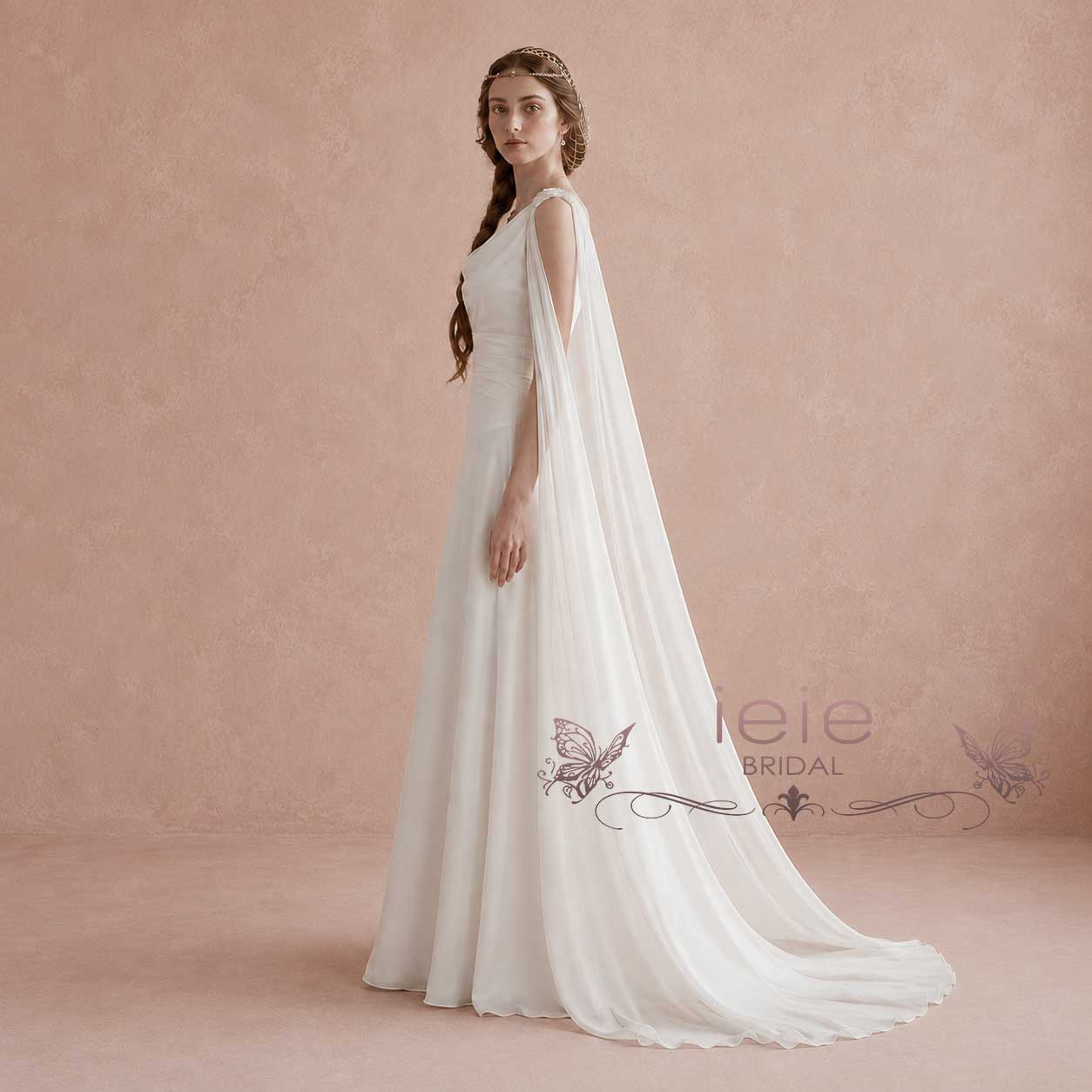 A woman modeling a floor-length white bridal gown with a flowing sheer cape train, standing against a warm beige studio backdrop. The dress has a fitted bodice and a long draped veil that trails behind her.