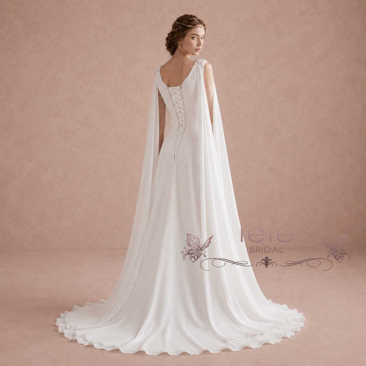 A woman modeling a white bridal gown with a draped cape-style back and lace-up open back, photographed against a warm beige studio backdrop. The dress is shown from the back, with a long train extending across the floor.