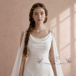 A woman wearing a white bridal gown with draped sheer fabric over the shoulders and a delicate headpiece, posed against a soft beige textured wall with warm window light casting shadows.