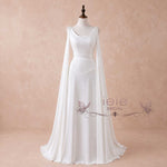 A white bridal gown displayed on a mannequin against a soft beige textured backdrop. The dress features a scoop neckline, draped cap sleeves, a fitted bodice, and a flowing skirt with a long sheer overskirt.