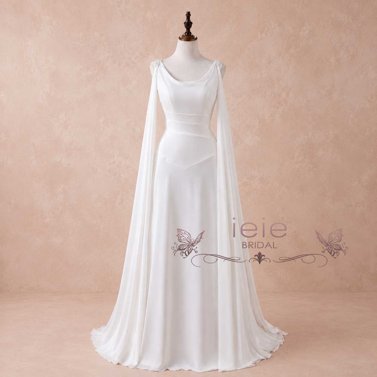 A white bridal gown displayed on a mannequin against a soft beige textured backdrop. The dress features a scoop neckline, draped cap sleeves, a fitted bodice, and a flowing skirt with a long sheer overskirt.