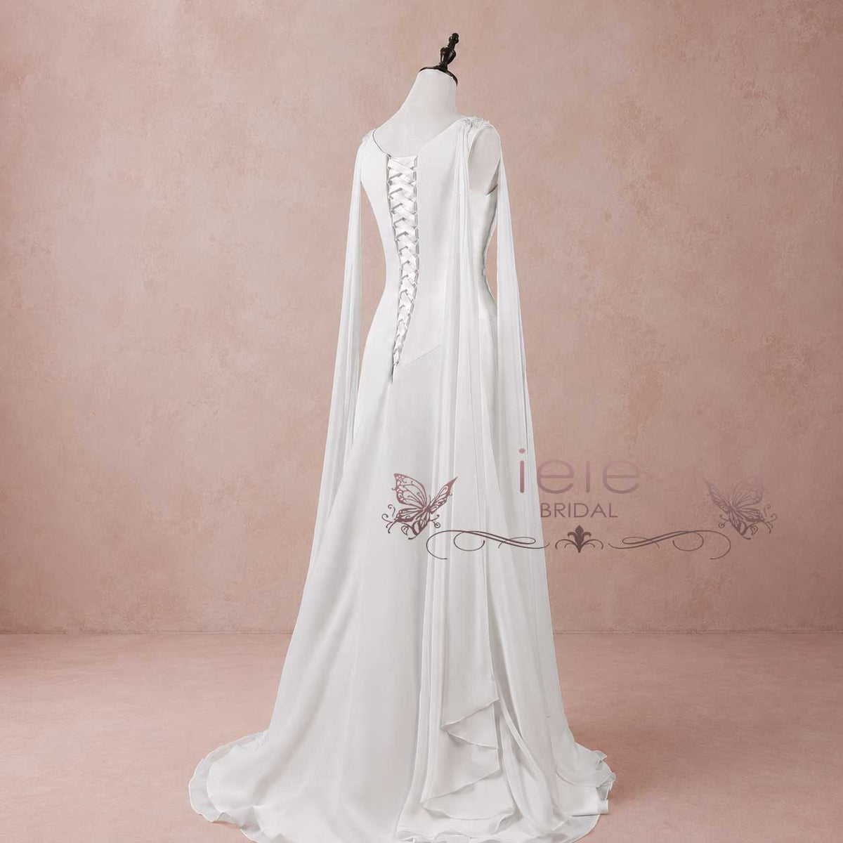 White bridal gown with a high-low train, long draped sleeves, and a lace-up back, displayed on a mannequin against a dusty pink studio backdrop.