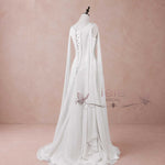 White bridal gown with a high-low train, long draped sleeves, and a lace-up back, displayed on a mannequin against a dusty pink studio backdrop.
